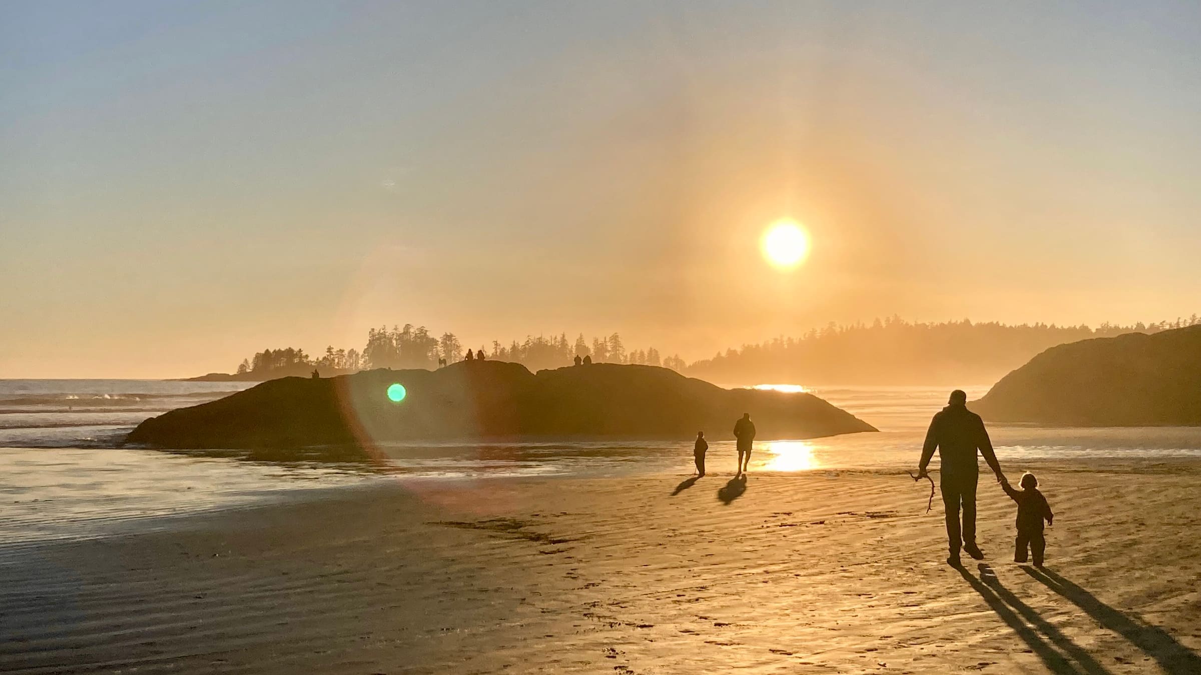 A parent and small child walking hand in hand along Long Beach at sunset, with rocky headlands and the Pacific behind them